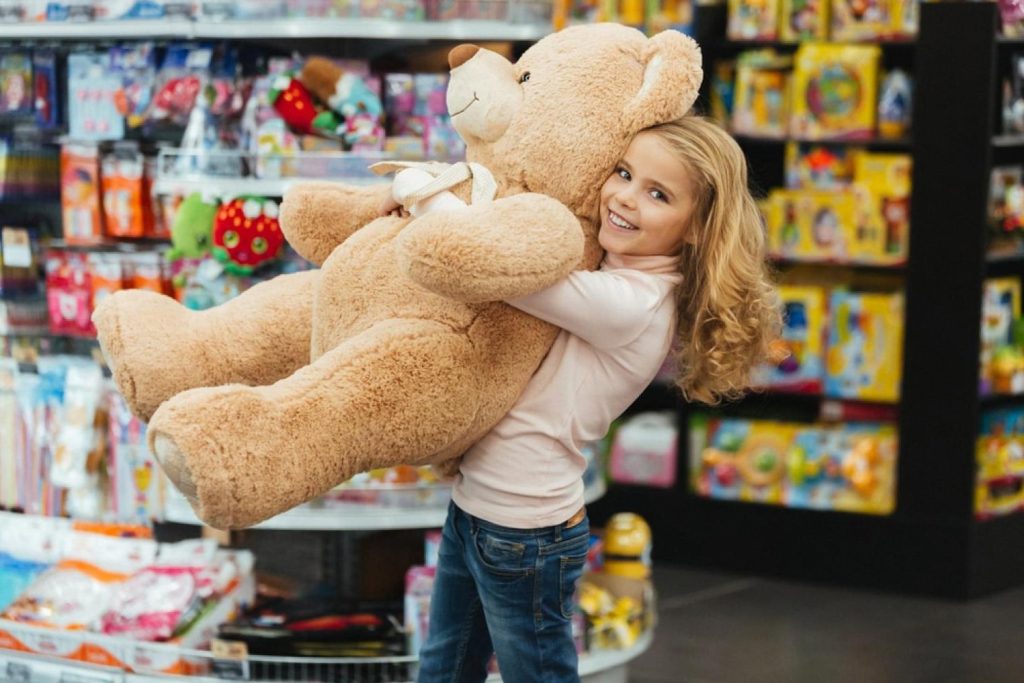 Smiling girl hugging a large teddy bear inside a colourful toy store